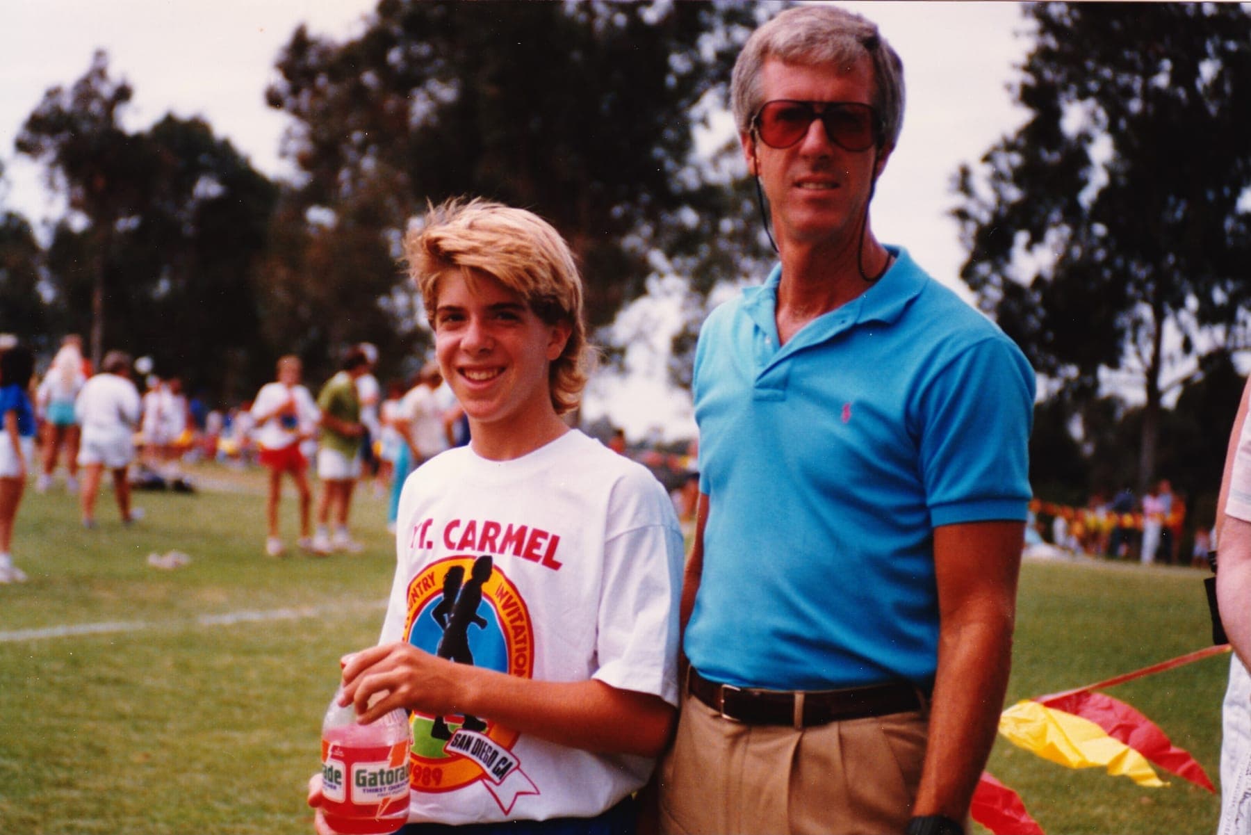 A teenage runner in a Mt. Carmel Cross Country Invitational shirt holding a Gatorade, standing with their dad on the grass at a 1989 meet in San Diego, spectators and course flags behind them.